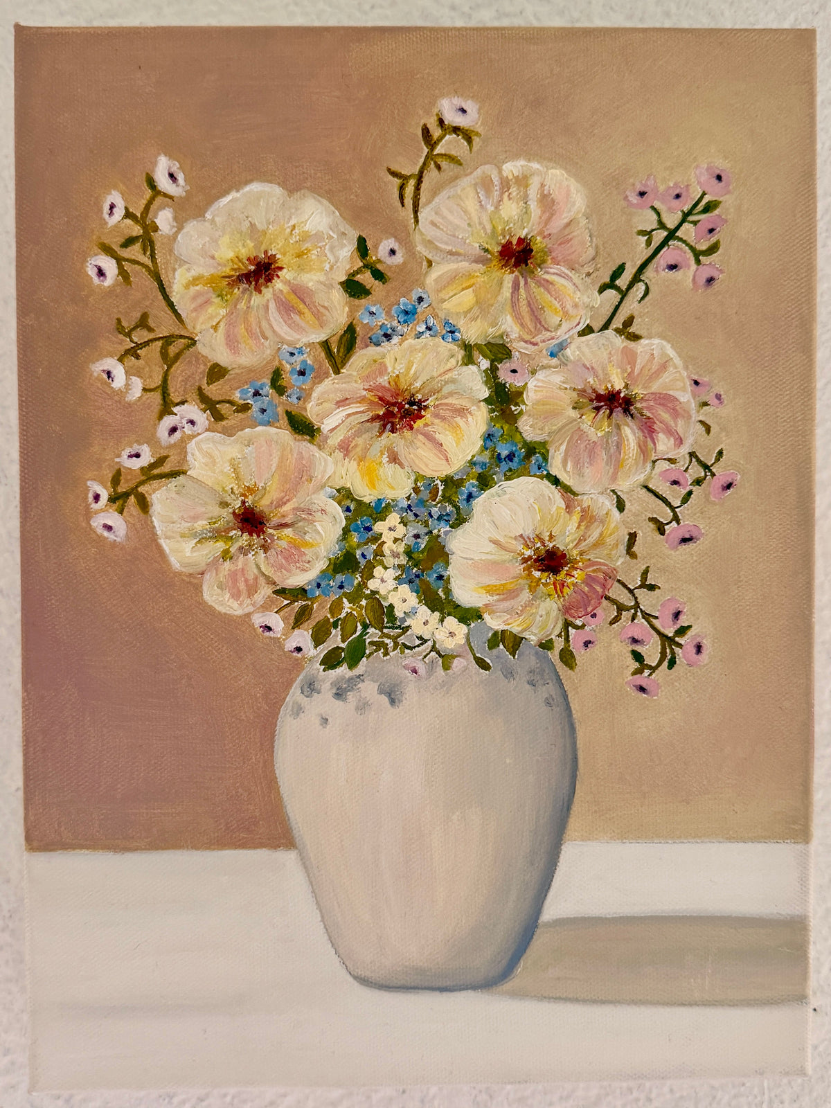 A still life painting of a bouquet of pale yellow and pink flowers with blue accents in a white vase, set against a warm brown background